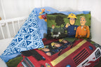 Ambiente-Aufnahme der Kinderbettwäsche „Feuerwehrmann Sam“ in einem weißen Bett; die Decke ist leicht umgeschlagen und zeigt die blaue Rückseite mit Sicherheits- und Warnsymbolen, während das Kissen am Kopfende platziert ist.