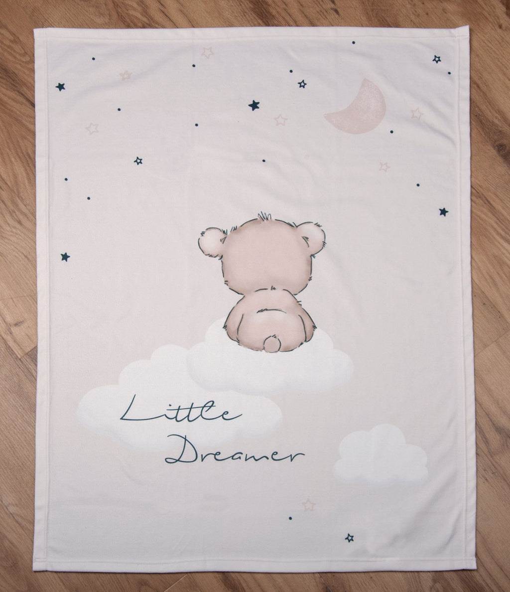 Draufsicht der Baby-Kuscheldecke komplett flach ausgebreitet auf hellem Holzboden; zeigt das zentrale Motiv des kleinen Bären, der von hinten auf einer Wolke sitzt, den Schriftzug „Little Dreamer“, Wolken, Sterne und eine Mondsichel.