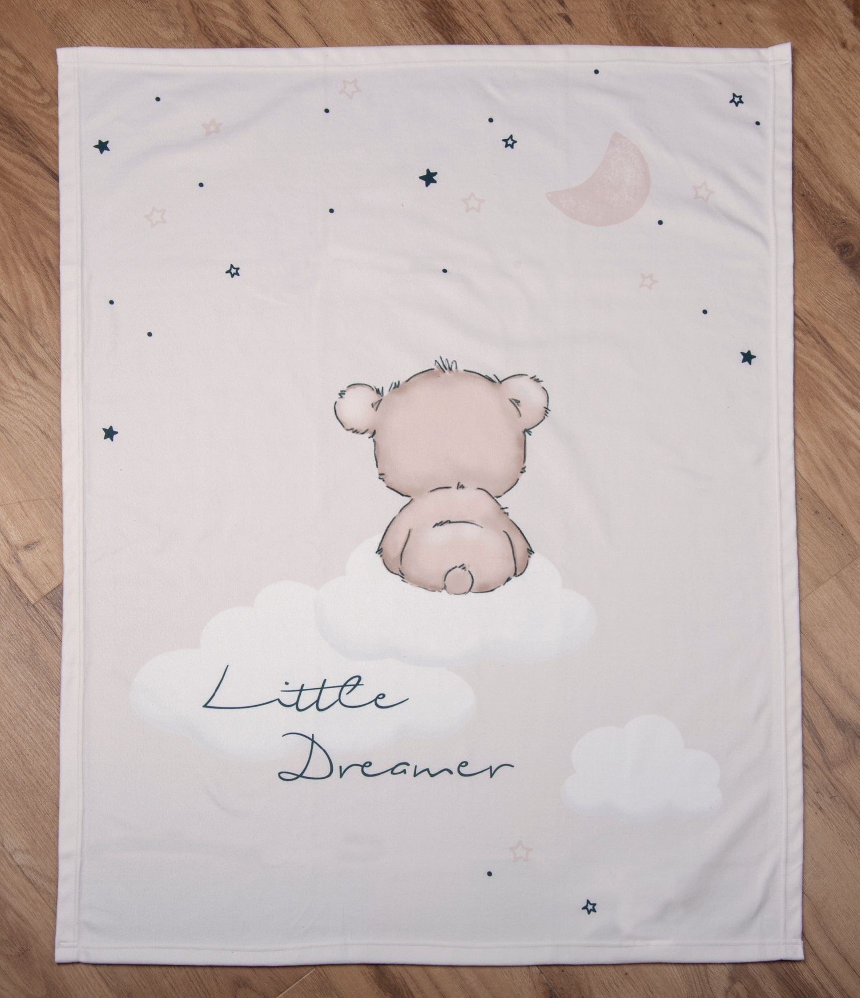 Draufsicht der Baby-Kuscheldecke komplett flach ausgebreitet auf hellem Holzboden; zeigt das zentrale Motiv des kleinen Bären, der von hinten auf einer Wolke sitzt, den Schriftzug „Little Dreamer“, Wolken, Sterne und eine Mondsichel.
