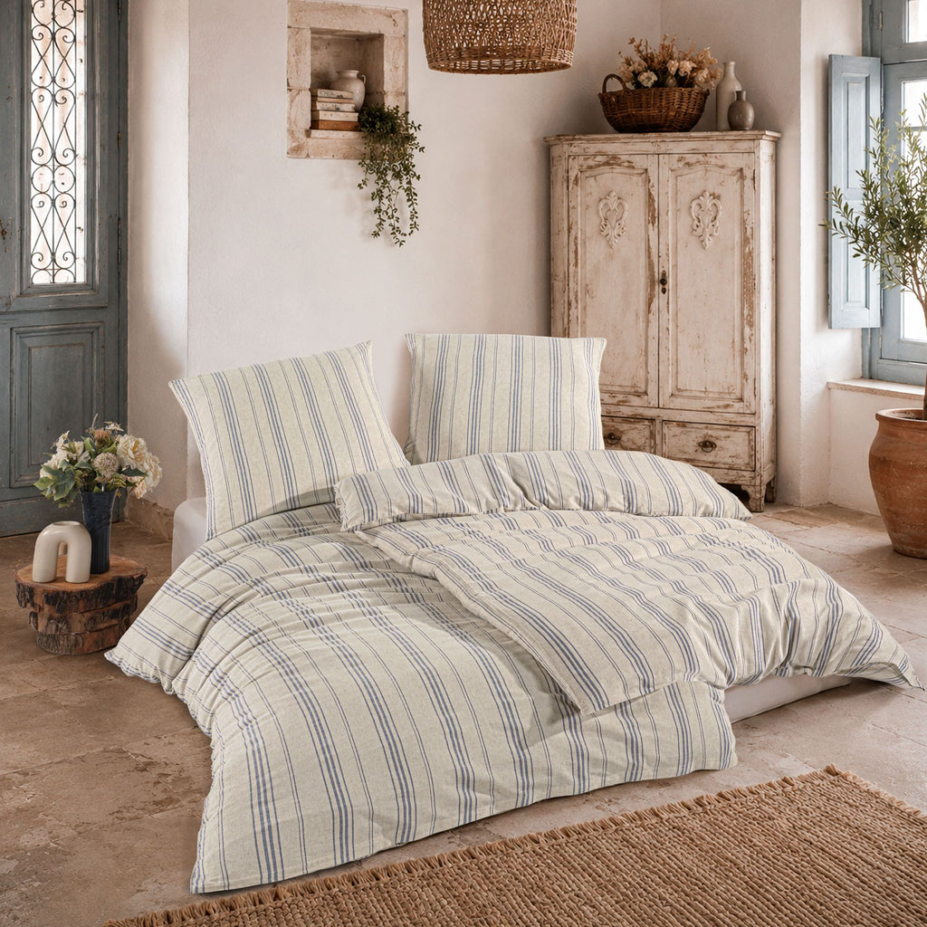 Bett mit blau-beiger Streifen-Bettwäsche auf Fliesenboden, flankiert von antikem Schrank und Pflanzen.