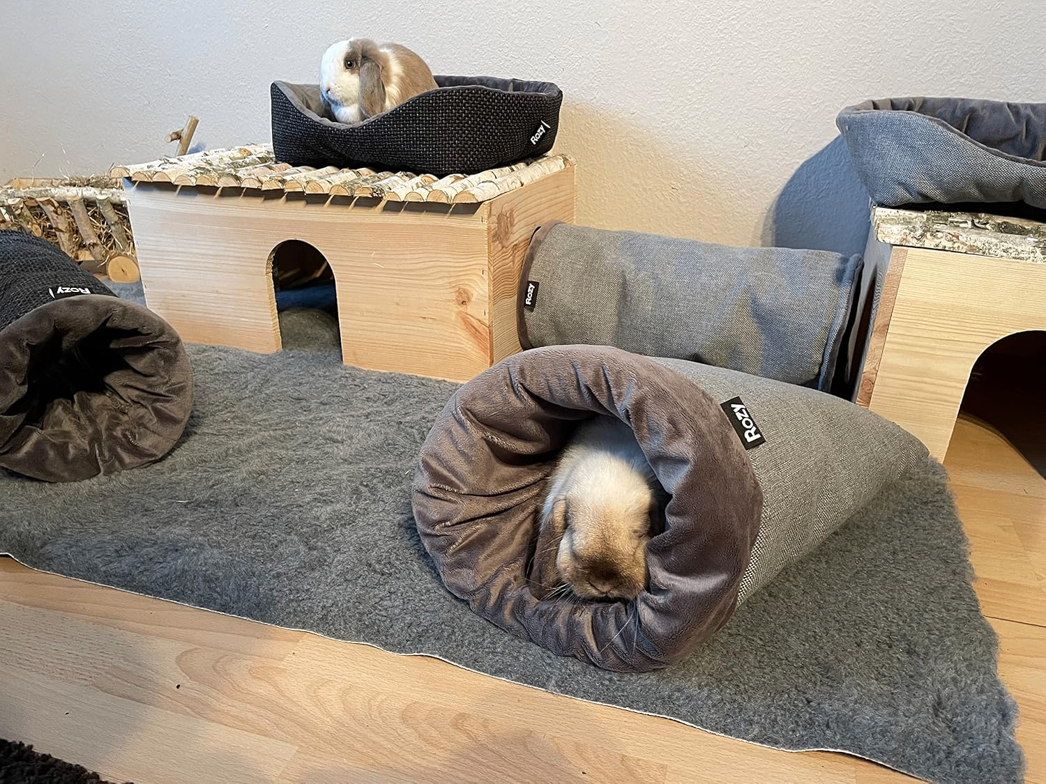 Kaninchengehege mit Holzmöbeln und grauem Teppich. Ein helles Kaninchen liegt entspannt im „Rozy“-Kuschelsack, ein anderes sitzt oben im separaten Bettchen.