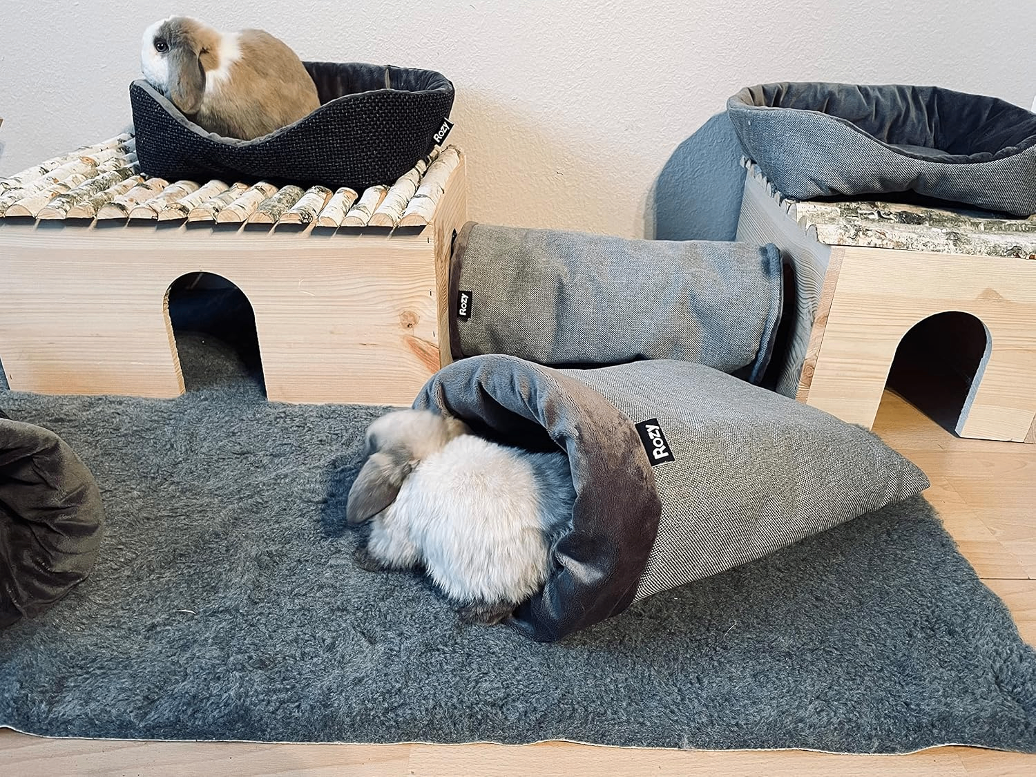 Nahaufnahme: Helles Kaninchen liegt tief im kegelförmigen „Rozy“-Kuschelsack auf grauem Teppich, während das dunklere, kleinere Kaninchen direkt vor der Öffnung ruht.