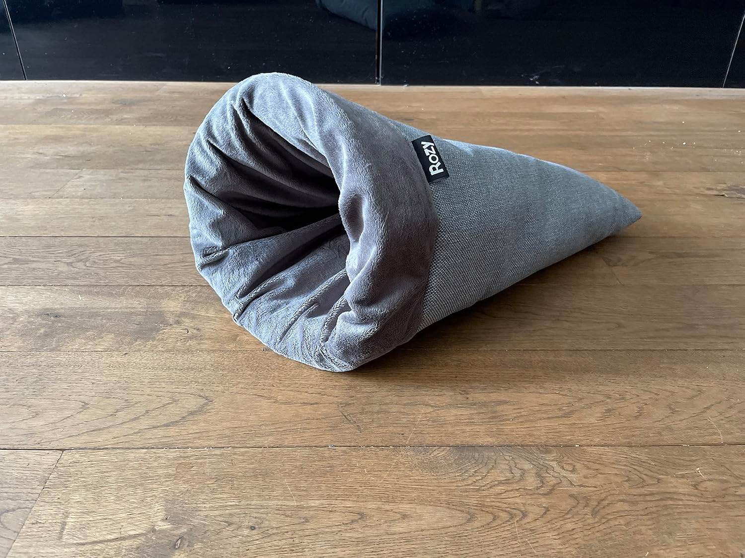 Produktansicht des grauen Kleintier-Kuschelsacks „Rozy“ auf einem Holzfußboden. Der kegelförmige Sack mit Plüschfutter liegt schräg auf dem Boden.