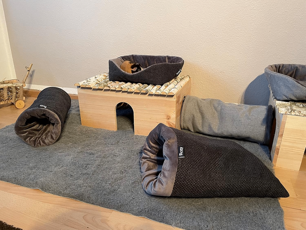 Meerschweinchengehege mit Zubehör. Ein dreifarbiges Meerschweinchen sitzt im Kuschelbett auf einem Holzhaus, davor liegen dunkle Kuschelsäcke.