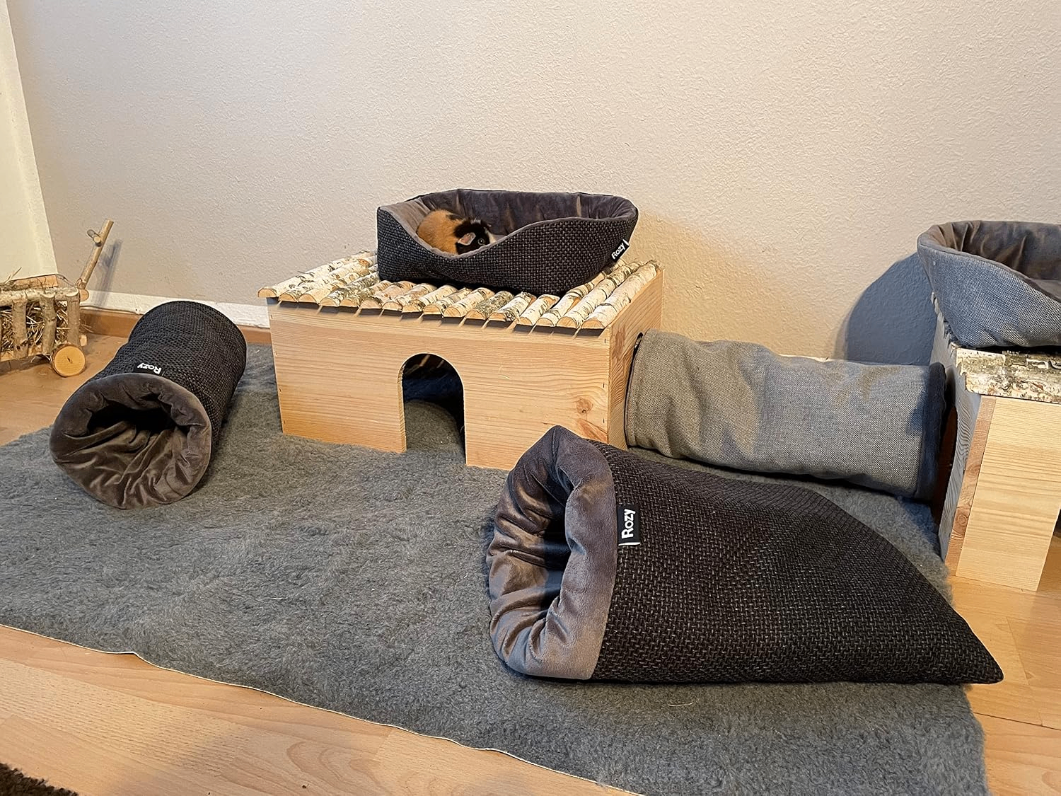 Meerschweinchengehege mit Zubehör. Ein dreifarbiges Meerschweinchen sitzt im Kuschelbett auf einem Holzhaus, davor liegen dunkle Kuschelsäcke.