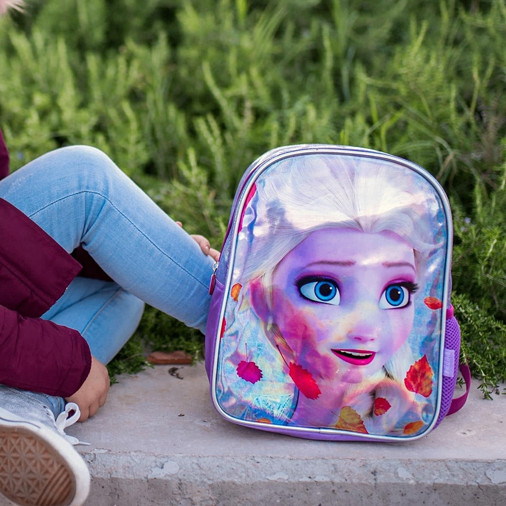 Ein Kind sitzt draußen neben dem lila Elsa-Rucksack, der auf einer Steinmauer steht. Im Hintergrund ist grünes Gebüsch zu sehen.
