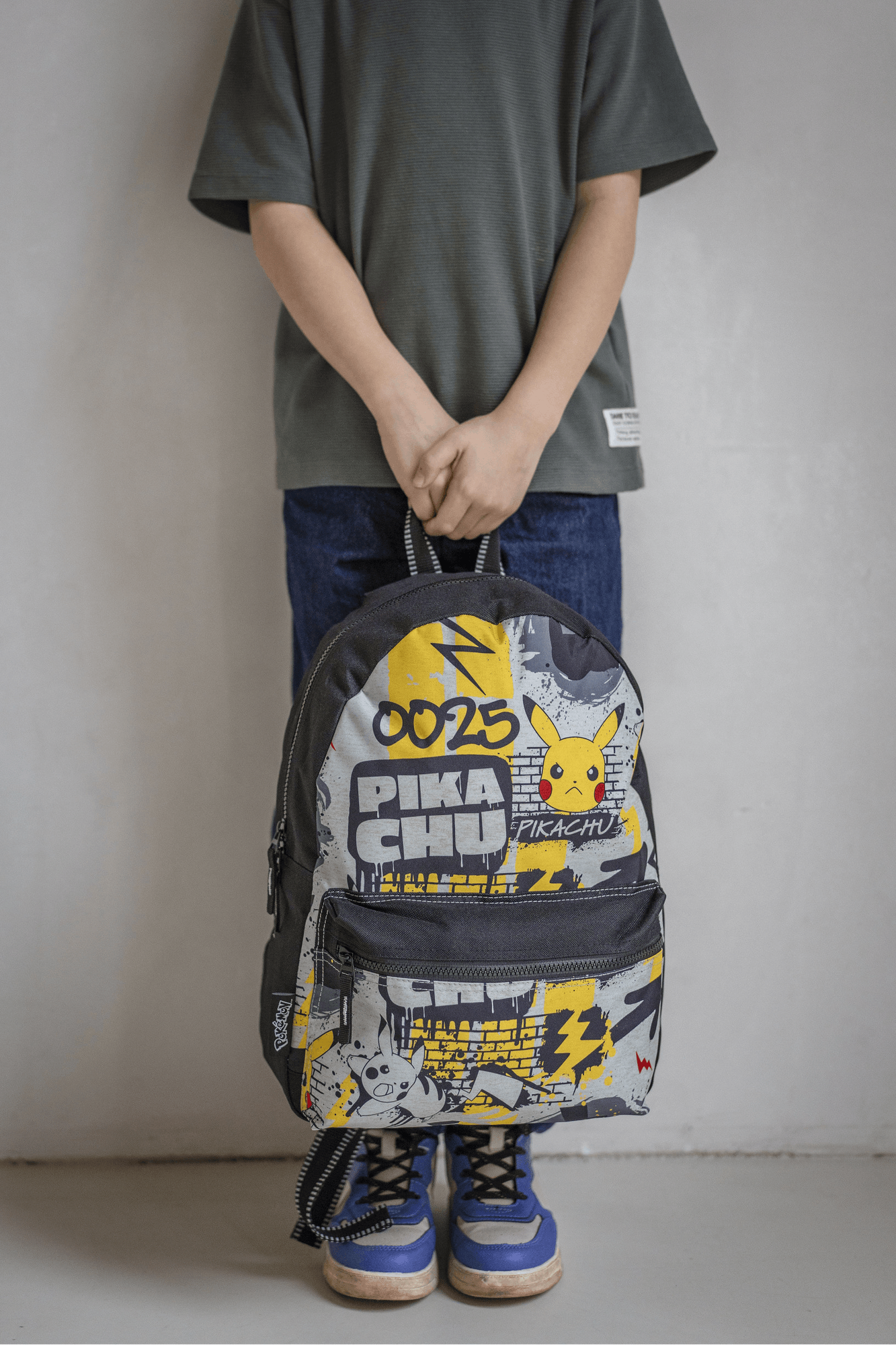 Kind hält den gelb-schwarzen Pokémon Pikachu Rucksack am oberen Tragegriff vor hellem Hintergrund.
