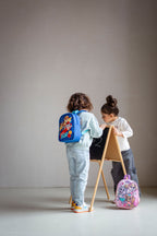 Kind mit blauem Paw Patrol Rucksack steht an einer kleinen Tafel, während ein weiteres Kind mit rosa Rucksack daneben steht.